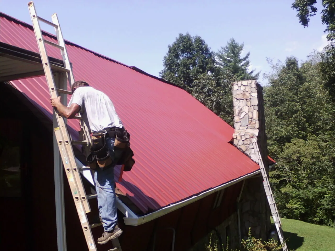 Roofer on a ladder beside a red metal roof and stone chimney — Soffit & Fascia Repair by Roofing Brothers in South Houston
